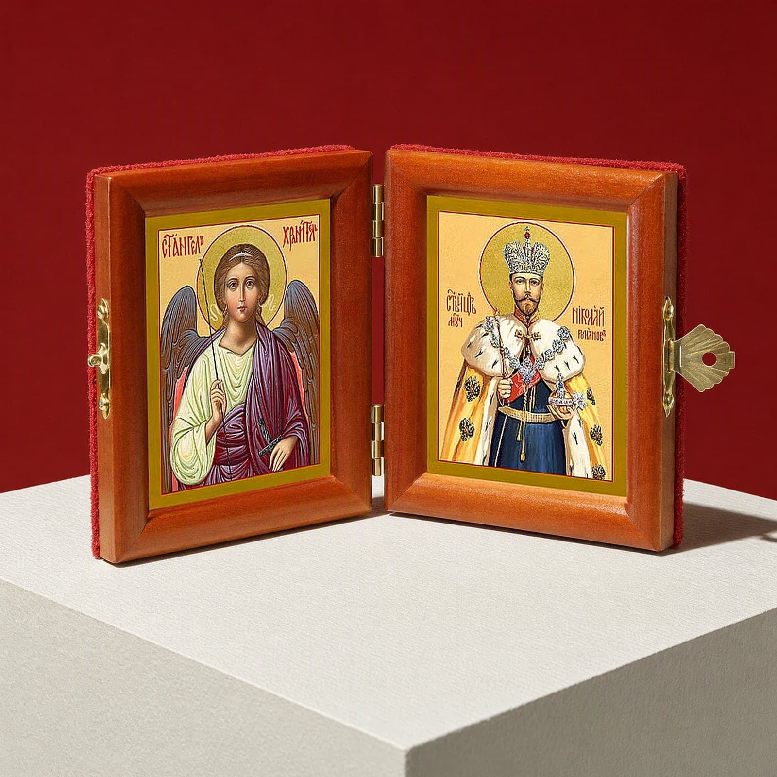 St Nicholas II (passion-bearer) & Guardian Angel name diptych seen from a 45-degree angle, half open on a white plinth against an oxblood backdrop, showing the frame depth and hinges.