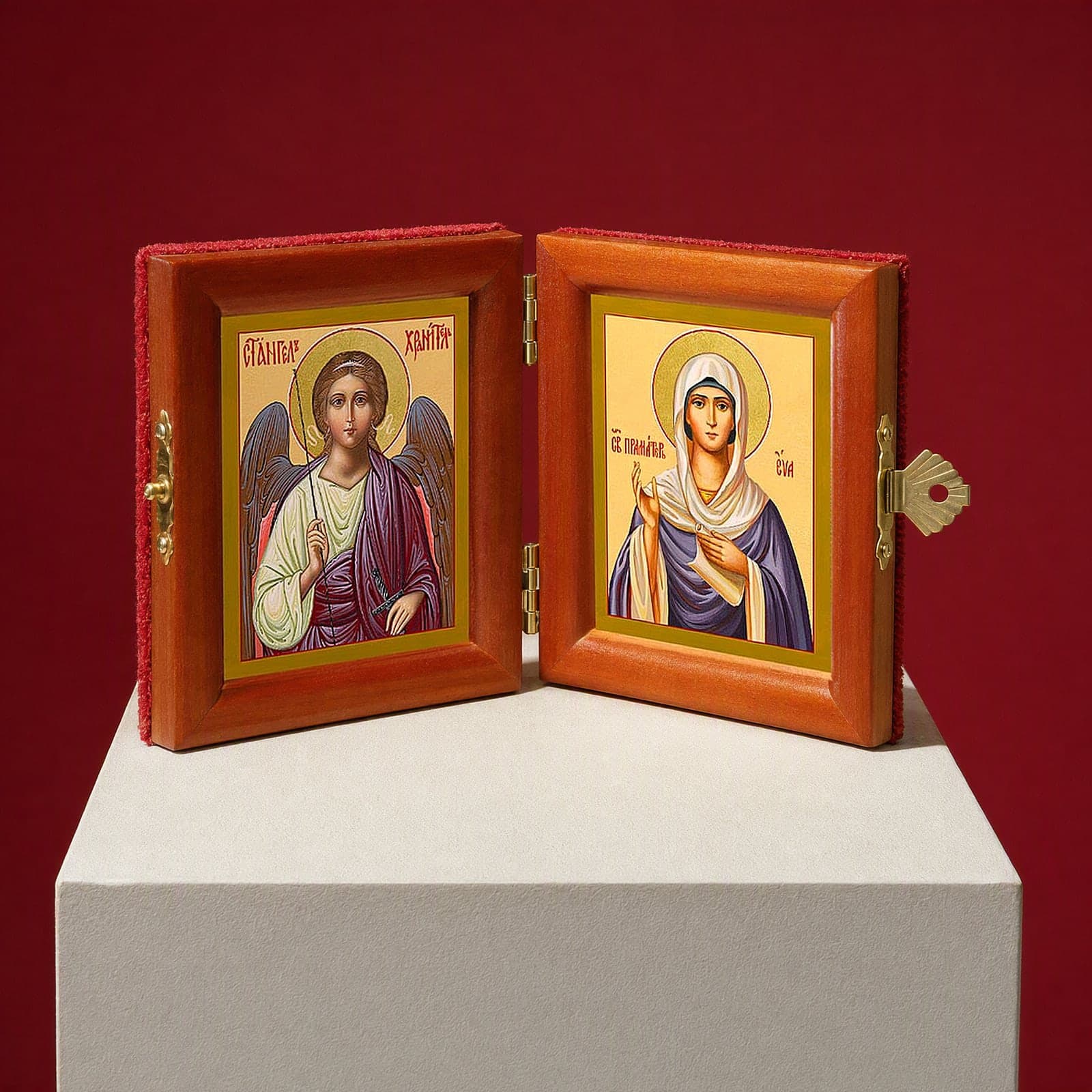 Holy Foremother Eve & Guardian Angel name diptych seen from a 45-degree angle, half open on a white plinth against an oxblood backdrop, showing the frame depth and hinges.