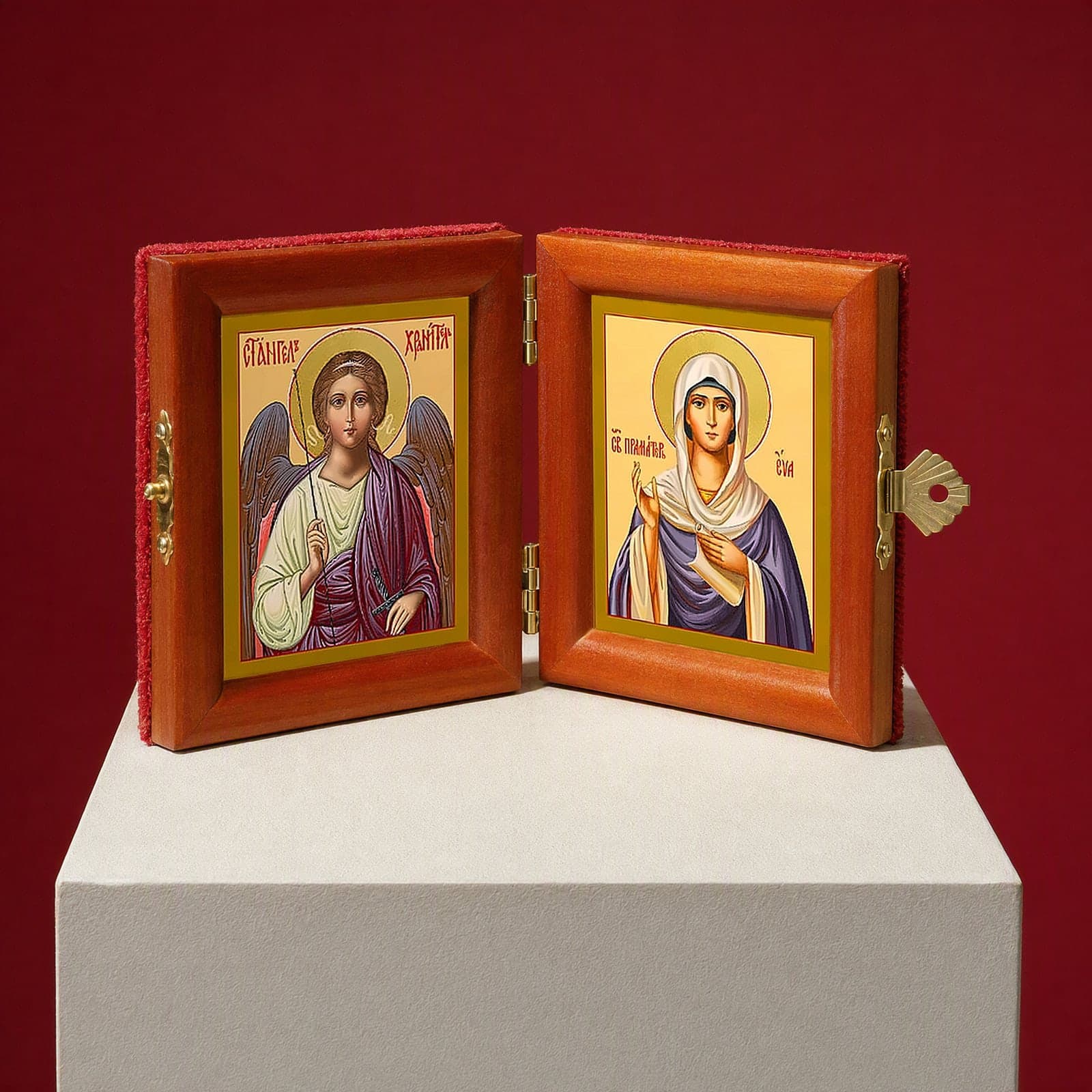 Holy Foremother Eve & Guardian Angel name diptych seen from a 45-degree angle, half open on a white plinth against an oxblood backdrop, showing the frame depth and hinges.