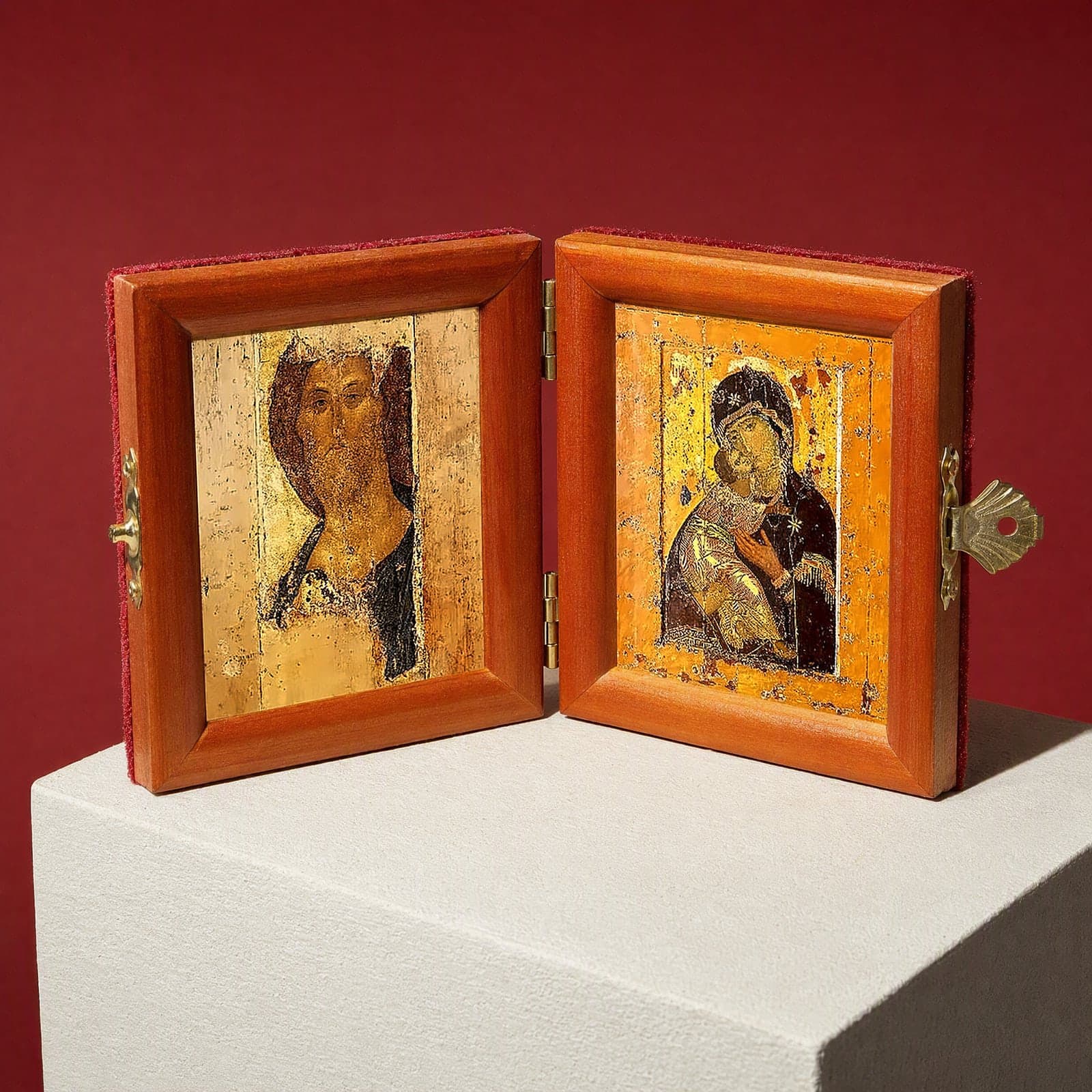 Christ (Zvenigorod Saviour) & Vladimir Mother of God travel diptych (Rublev era) seen from a 45-degree angle, half open on a white plinth against an oxblood backdrop, showing the frame depth and hinges.