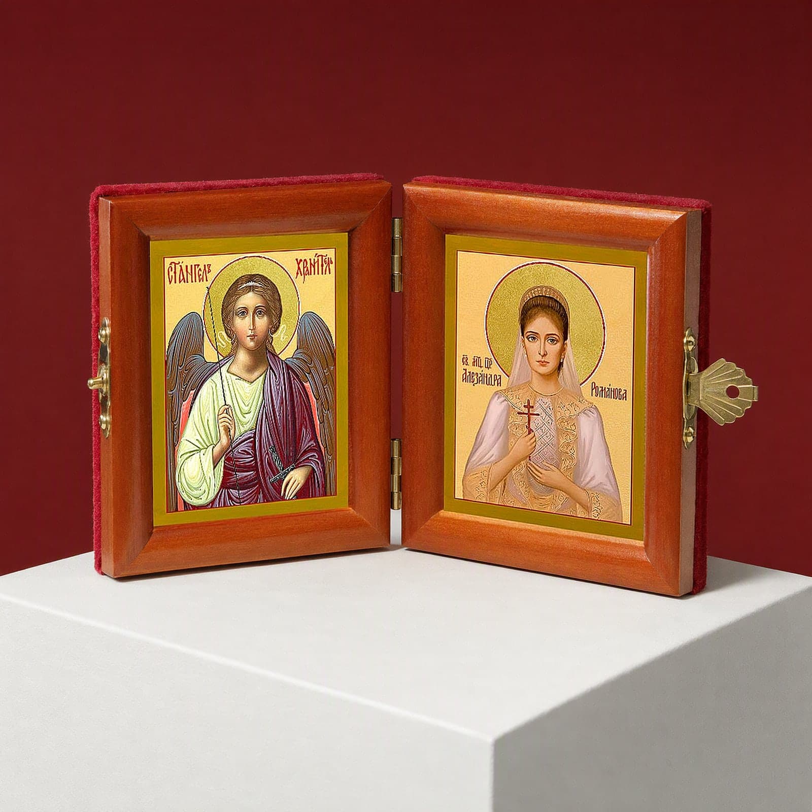 St Alexandra (passion-bearer Empress) & Guardian Angel name diptych seen from a 45-degree angle, half open on a white plinth against an oxblood backdrop, showing the frame depth and hinges.