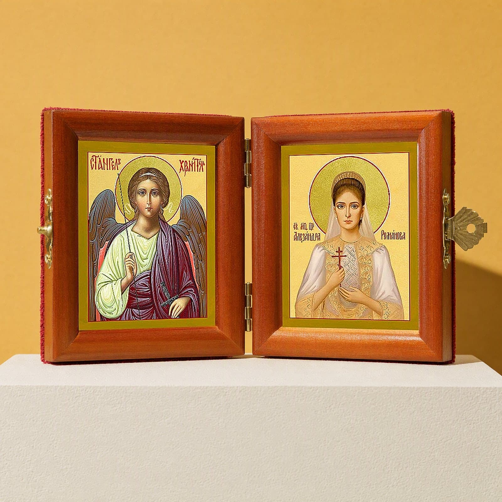 St Alexandra (passion-bearer Empress) & Guardian Angel name diptych, open: folding diptych of the Guardian Angel and St Alexandra the passion-bearer, Empress in honey pine frames with brass hinges and clasp, on a white plinth against an ochre backdrop.