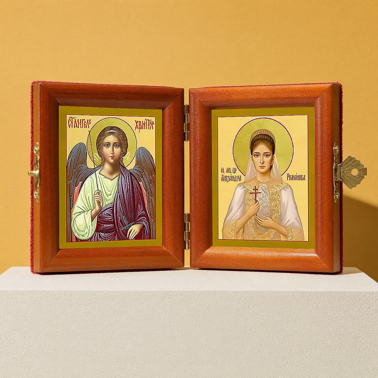 St Alexandra (passion-bearer Empress) & Guardian Angel name diptych, open: folding diptych of the Guardian Angel and St Alexandra the passion-bearer, Empress in honey pine frames with brass hinges and clasp, on a white plinth against an ochre backdrop.