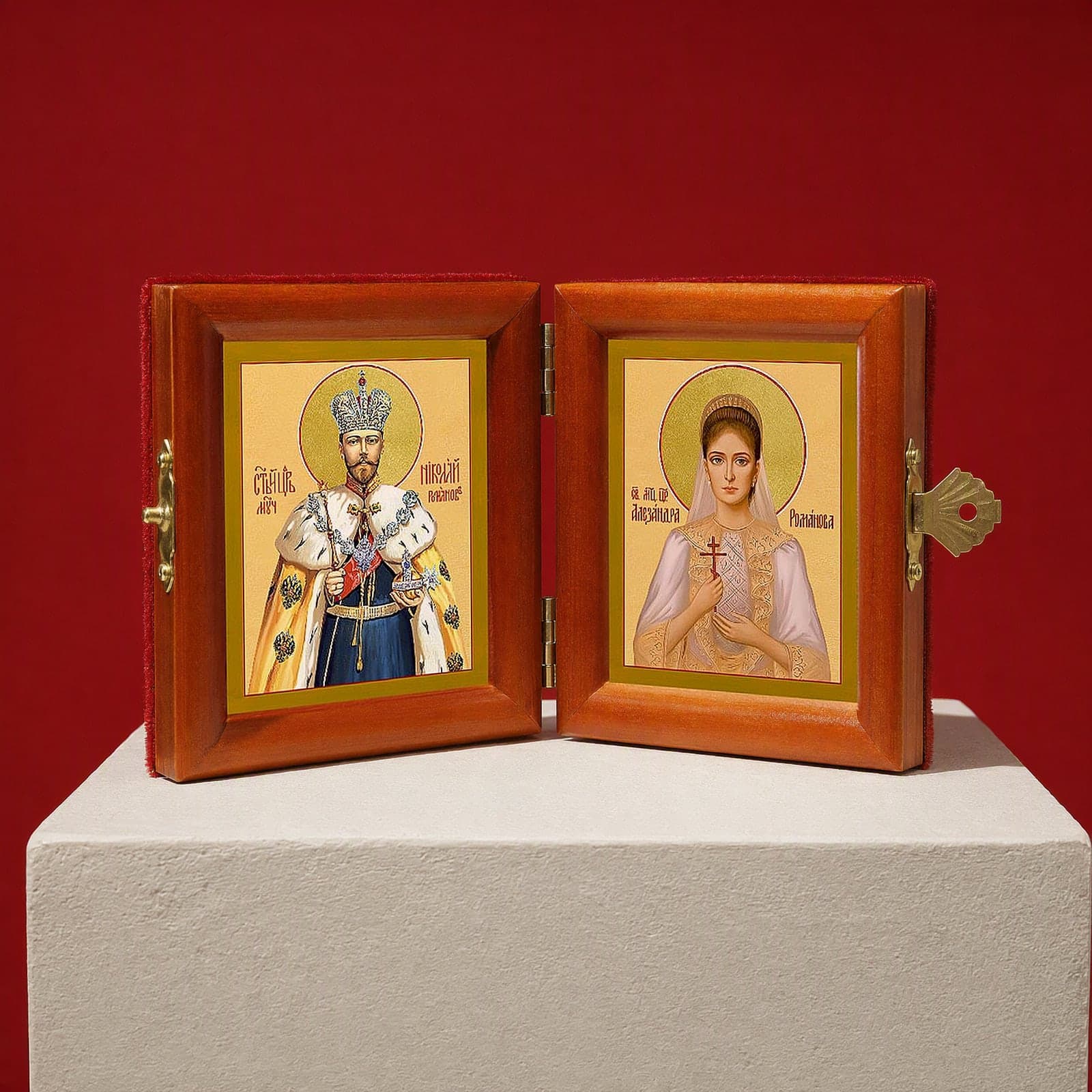 Sts Nicholas II & Alexandra (Royal Passion-Bearers) diptych seen from a 45-degree angle, half open on a white plinth against an oxblood backdrop, showing the frame depth and hinges.