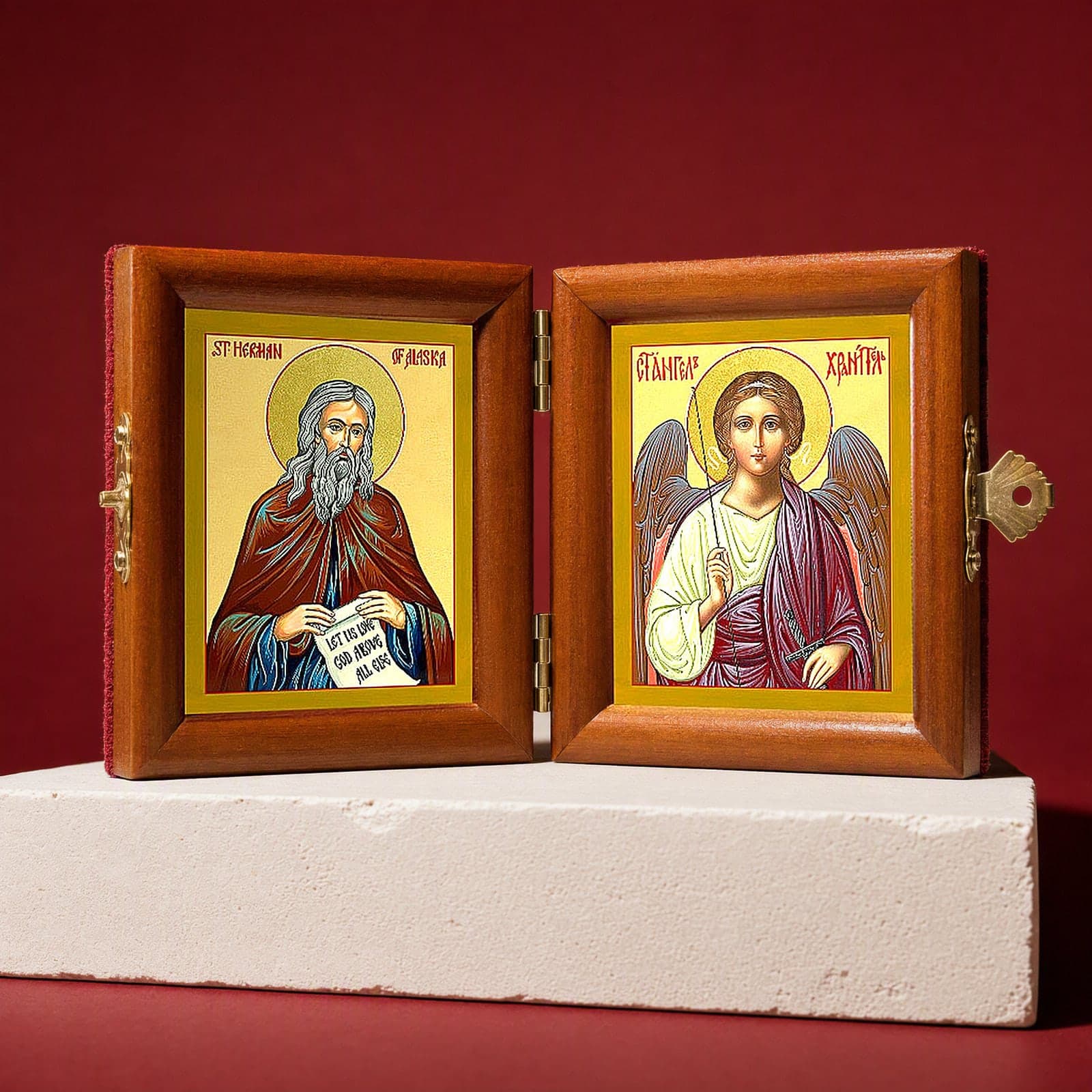 St Herman of Alaska & Guardian Angel name diptych seen from a 45-degree angle, half open on a white plinth against an oxblood backdrop, showing the frame depth and hinges.