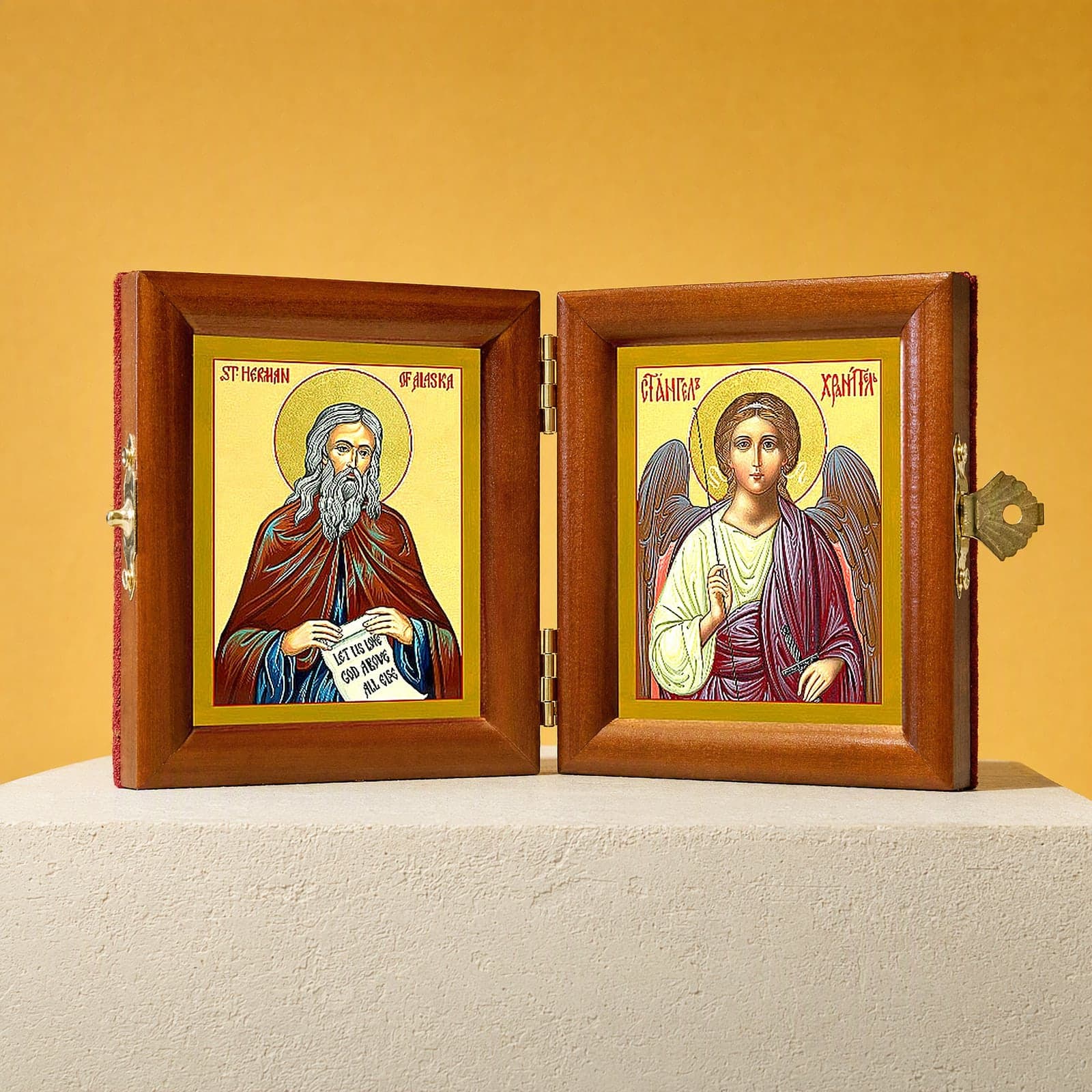 St Herman of Alaska & Guardian Angel name diptych, open: folding diptych of St Herman of Alaska holding a scroll and the Guardian Angel in honey pine frames with brass hinges and clasp, on a white plinth against an ochre backdrop.