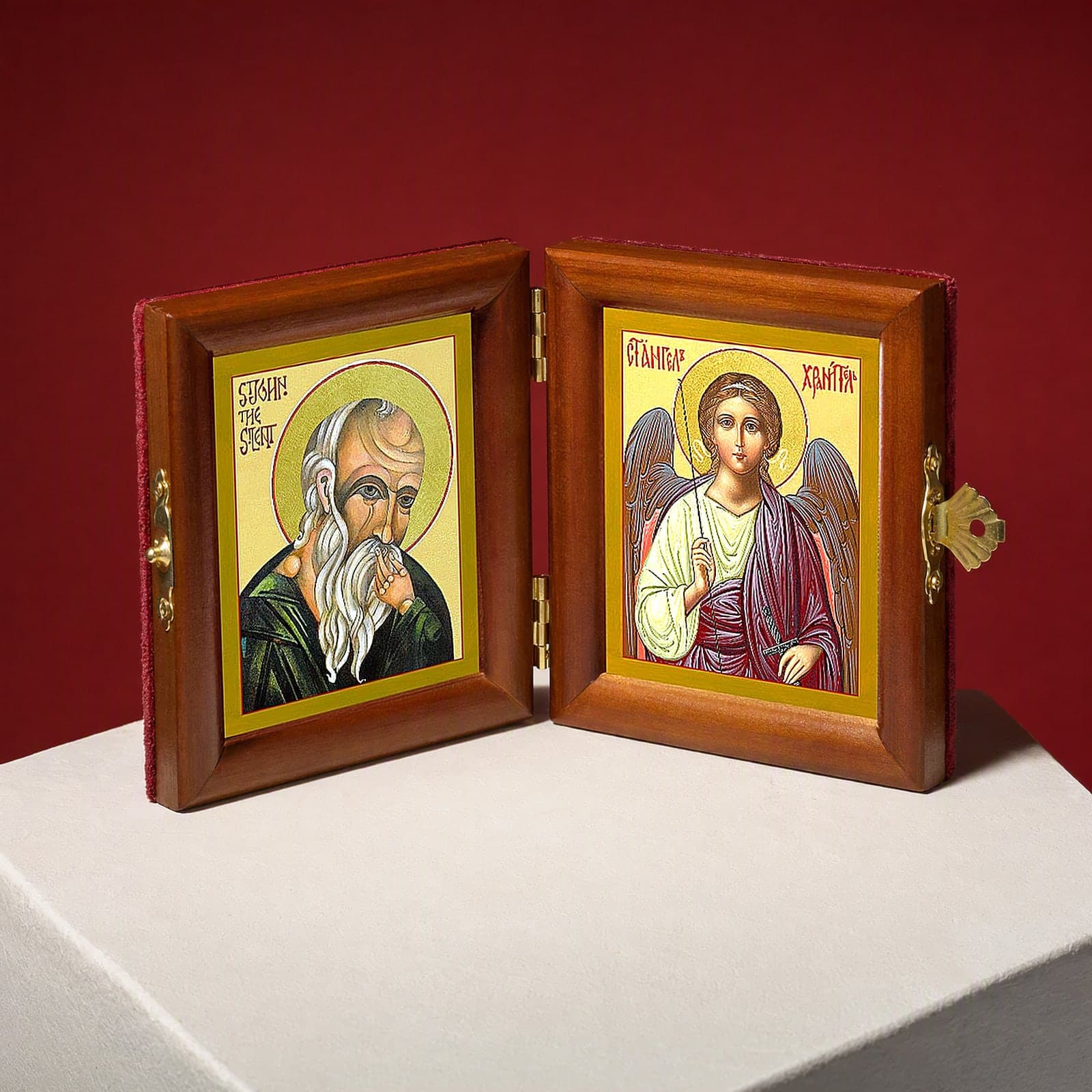 St John the Silent & Guardian Angel name diptych seen from a 45-degree angle, half open on a white plinth against an oxblood backdrop, showing the frame depth and hinges.