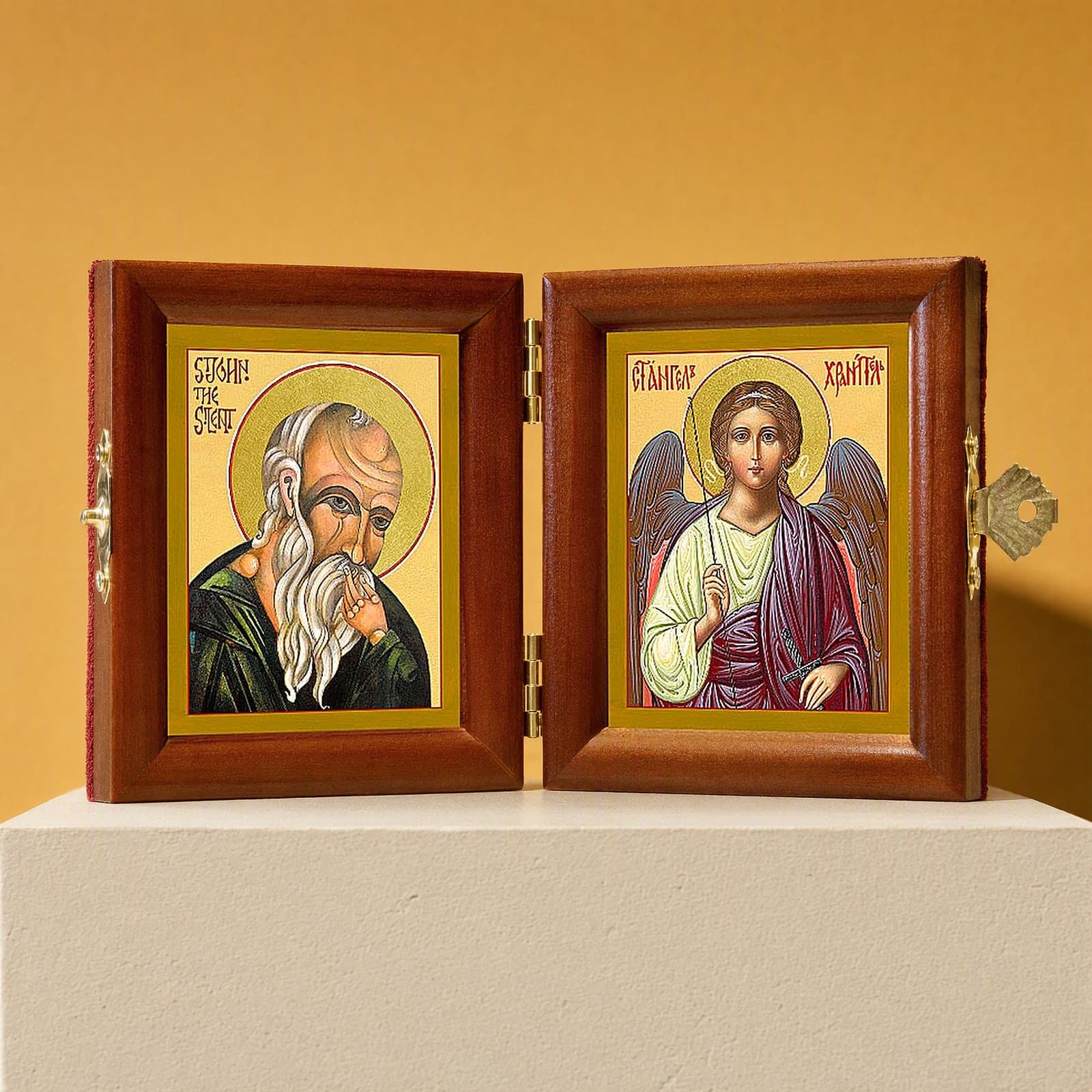 St John the Silent & Guardian Angel name diptych, open: folding diptych of St John the Silent and the Guardian Angel in honey pine frames with brass hinges and clasp, on a white plinth against an ochre backdrop.