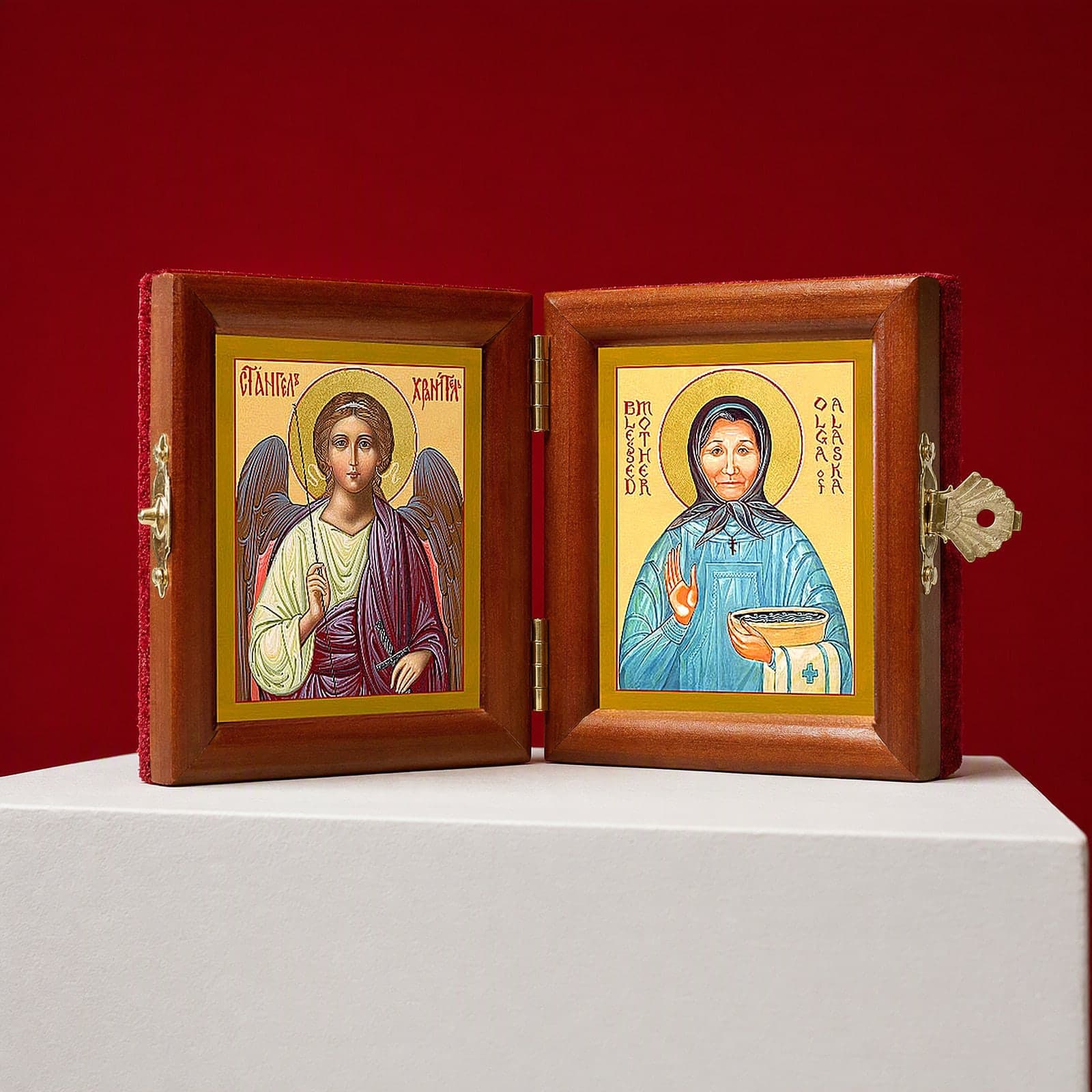 St Olga of Alaska & Guardian Angel name diptych seen from a 45-degree angle, half open on a white plinth against an oxblood backdrop, showing the frame depth and hinges.