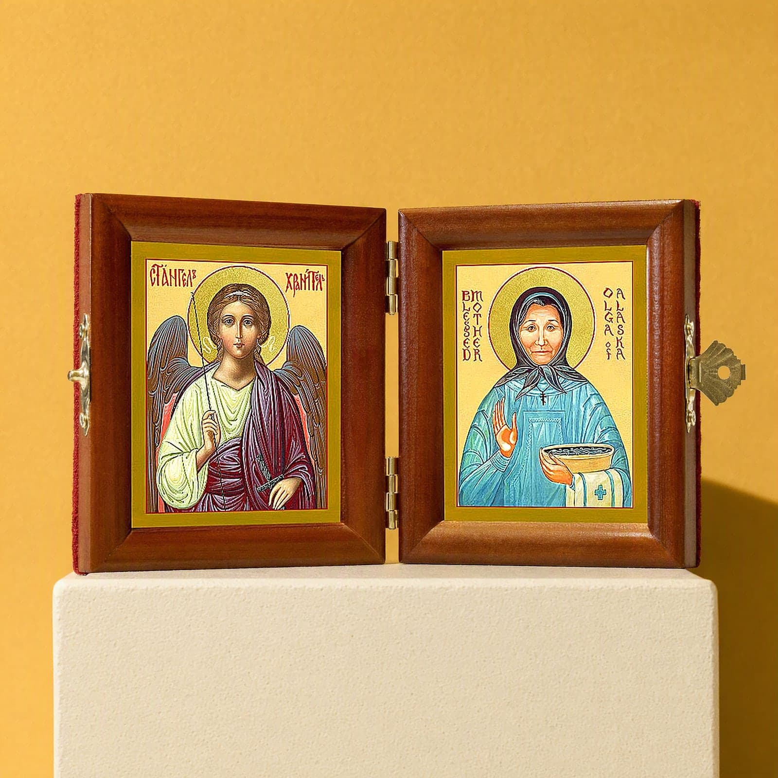 St Olga of Alaska & Guardian Angel name diptych, open: folding diptych of the Guardian Angel and St Olga of Alaska in honey pine frames with brass hinges and clasp, on a white plinth against an ochre backdrop.