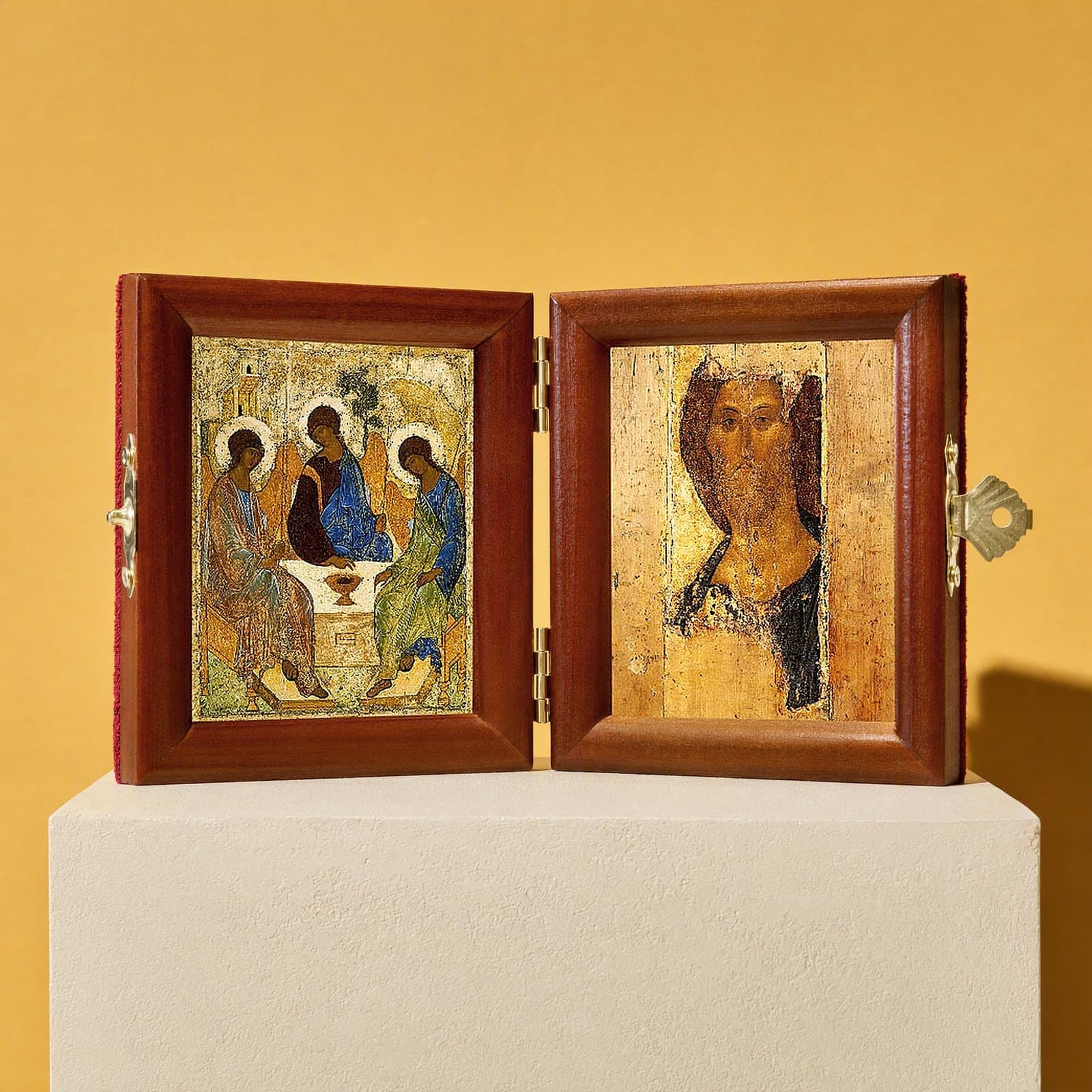 Holy Trinity (Rublev) & Christ (Zvenigorod Saviour) travel diptych, open: folding diptych of the Holy Trinity of Rublev, three angels at a table and Christ in the Rublev manner (the Zvenigorod Saviour) in honey pine frames with brass hinges and clasp, on a white plinth against an ochre backdrop.